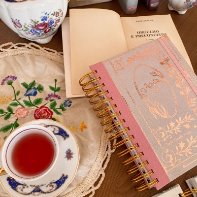 Agenda rosa 2026, chávena com chá, jarra decorativa, livro aberto e toalha bordada sobre mesa de madeira