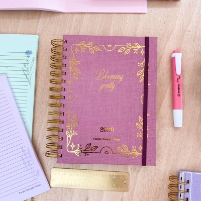 Caderno espiral roxo com detalhes dourados e elástico na mesa com caneta e réguas.