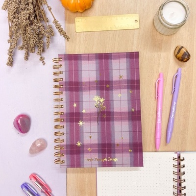 Caderno espiral xadrez roxo e rosa com canetas, régua, vela, pedras e abóbora decorativa num fundo de madeira e lilás