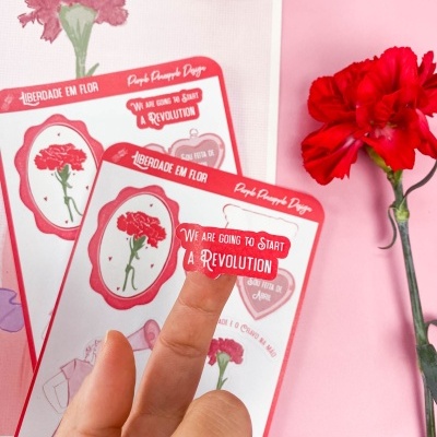 Adesivos com cravo vermelho e frases sobre fundo rosa com uma mão segurando um adesivo e flor real vermelha ao lado