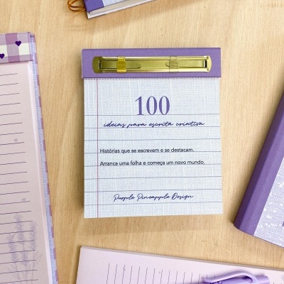 Bloco de notas roxo com prendedor dourado e texto em português, rodeado por materiais de escrita lilás numa mesa de madeira.
