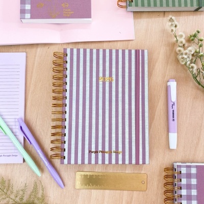 Conjunto de cadernos e agendas espiraladas listradas em roxo, lilás e verde com acessórios e flores sobre mesa de madeira clara