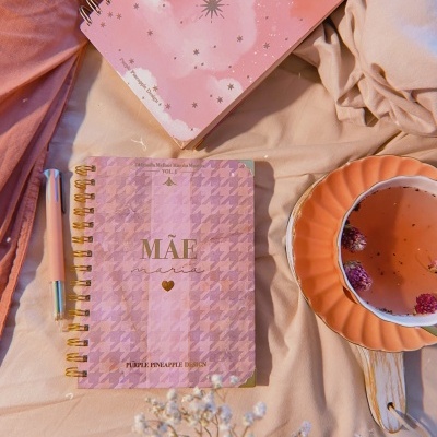 Dois cadernos rosas com mensagens, caneta, chá e flores sobre tecido cor-de-rosa e bege.