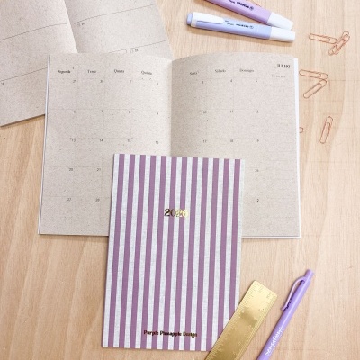 Agenda 2026 riscada lilás e branca com calendário, marcadores fluorescentes, caneta lilás e régua dourada sobre madeira