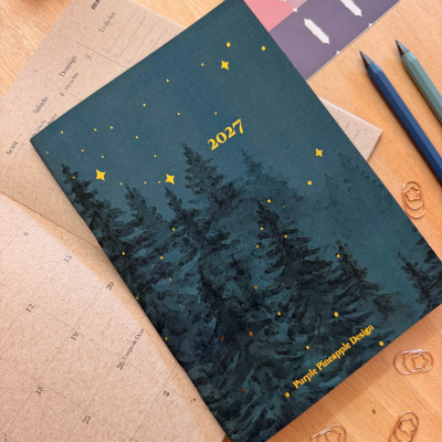 Calendário 2027 com capa azul e decoração de árvores e estrelas, sobre mesa de madeira com canetas e etiquetas adesivas
