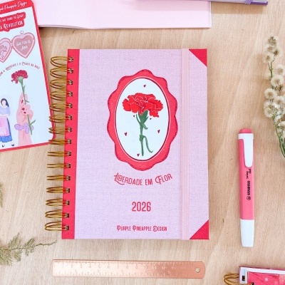 Caderno 2026 com capa rosa clara, ilustração de cravo vermelho e texto 'LIBERDADE EM FLOR', aneis dourados e banda elástica vermelha.
