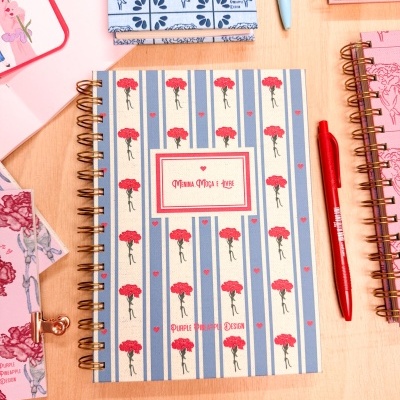 Caderno com capa azul e branca, flores vermelhas, caneta vermelha, sobre mesa de madeira