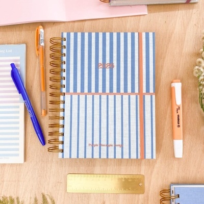 Agenda 2024 capa azul riscas brancas e laranjas com textos dourados e espiral dourada sobre mesa de madeira com acessórios