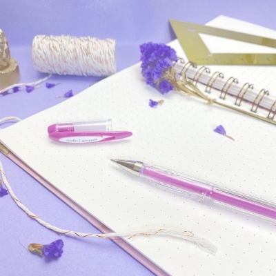 Caderno com caneta rosa, régua dourada, fio e flores secas lilás sobre fundo lilás