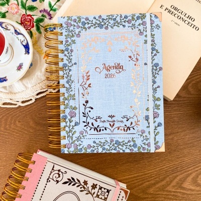 Dois cadernos com espiral dourado e capas florais, um azul claro e outro rosa claro, ambos com texto Agenda 2026.