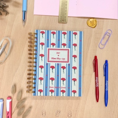 Agenda decorada com flores vermelhas e listas azuis junto a canetas coloridas, régua dourada e clips sobre mesa de madeira