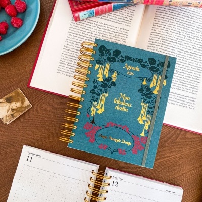 Agenda azul decorada com flores e texto dourado em mesa com livros e framboesas