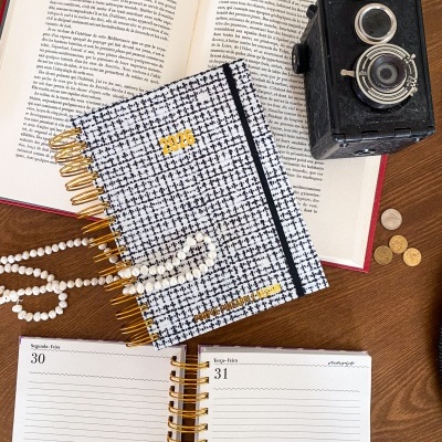 Agendas 2026 com capa padrão xadrez preto e branco, elástico preto, livro aberto em francês, câmera fotográfica preta vintage, moedas e colar de pérolas numa mesa de madeira
