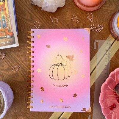 Caderno rosa degradê com abóbora e folhas douradas sobre mesa de madeira com velas, pedra e objetos diversos