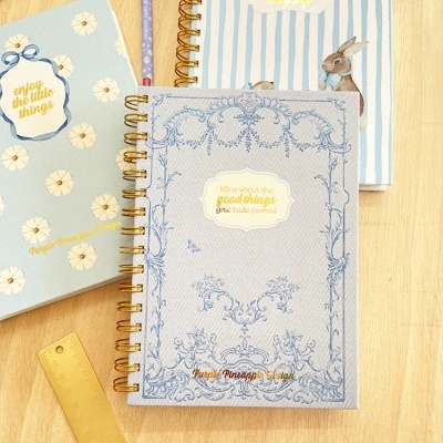 Três cadernos pastéis com espiral dourada e textos em inglês, régua dourada e lápis num fundo de madeira.