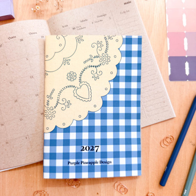 Agenda 2027 com padrão floral e de quadrados azul e branco sobre mesa de madeira com material de escritório