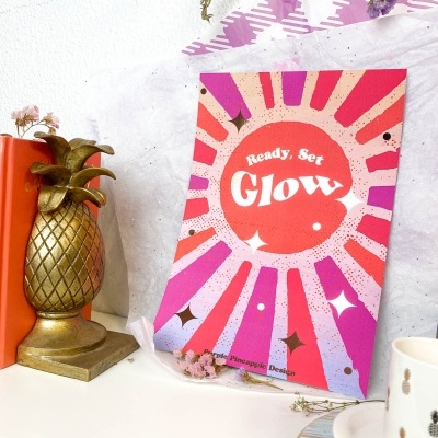 Cartaz colorido com texto 'Ready, Set Glow' numa mesa com decoração diversificada