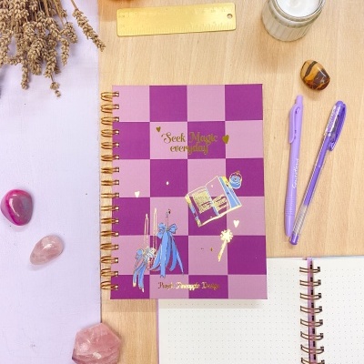 Caderno roxo quadriculado com decoração dourada, canetas lilás, régua, cristais e vela sobre mesa