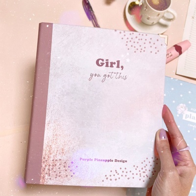 Caderno branco com texto 'Girl, you got this' e lombada rosa numa mesa com caneta, café, flores e outro caderno.