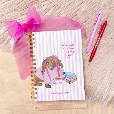 Caderno com capa ilustrada de cão castanho com laços rosa e livros, fita rosa, canetas e régua sobre madeira e tapete felpudo branco