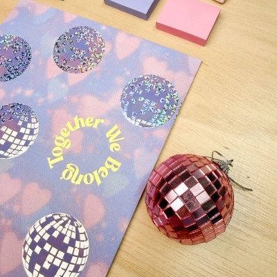 Bloco de notas, marcador STABILO, papel decorativo com texto Together We Belong e esfera decorativa vermelha em superfície de madeira.