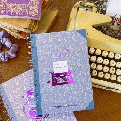 Caderno azul com máquina de escrever roxa e texto, ao lado de máquina de escrever amarela e livro roxo