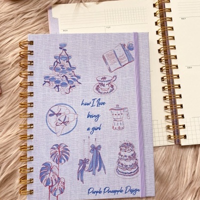 Agenda lilás com ilustrações e texto, espiral dourada e elástico, sobre superfície peluda rosa clara