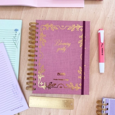 Caderno roxo com detalhes dourados, régua, caneta rosa em mesa de madeira.