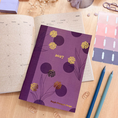 Agenda 2027 com capa roxa decorada com flores douradas e roxas sobre mesa com materiais de escritório
