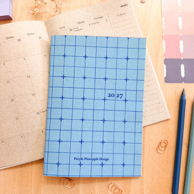 Agenda 2027 azul quadriculada com texto Purple Pineapple Design sobre mesa de madeira