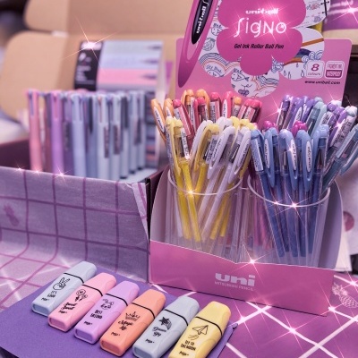 Esferográficas de gel uni-ball Signo em suporte rosa com várias cores pastéis e borrachas coloridas sobre tecido xadrez lilás e roxo.