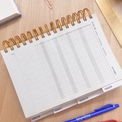 Calendário mensal de mesa com abas etiquetadas, canetas, clips e régua dourada sobre mesa de madeira.