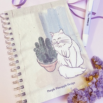Caderno com capa de tecido com desenho de gato branco e cacto, caneta e flores secas