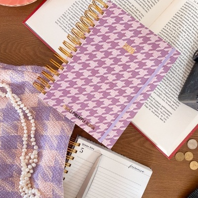 Caderno com capa pied-de-poule rosa e lilás sobre livro aberto, tecido e colar de pérolas próximo.