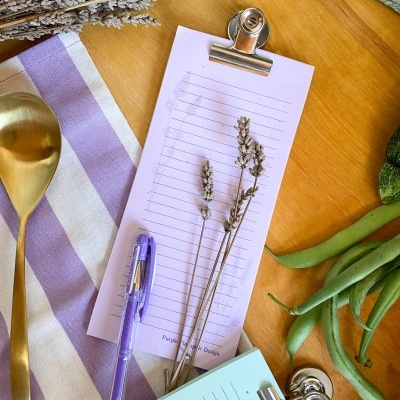 Mesa com utensílios de cozinha, pranchetas e itens decorativos em tons lilás, dourado e verde.