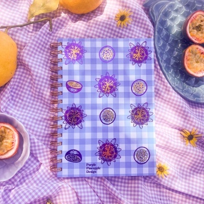 Caderno com capa xadrez lilás e ilustrações de maracujás sobre tecido xadrez lilás com frutas e flores