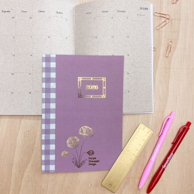 Agenda 2026 roxa com padrão xadrez e ilustração, sobre mesa com calendário e material de escritório