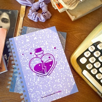 Caderno roxo com desenho de coração partido em frasco sobre mesa com máquina de escrever e outros objetos