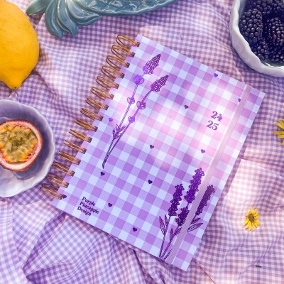 Caderno com capa xadrez roxa e branca com ilustrações de lavanda sobre pano xadrez, rodeado de frutas e flores amarelas