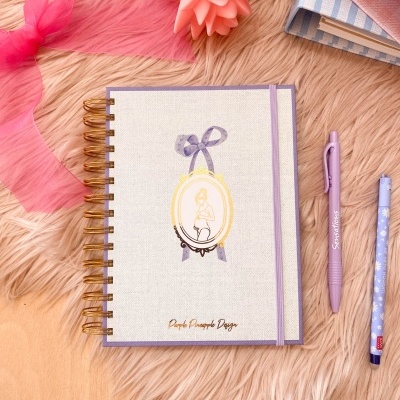 Caderno espiral branco e lilás com ilustração e texto, sobre pelo cor de creme com canetas, fita rosa e livros