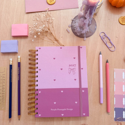 Planner 2027 rosa e roxo com selo de cera, lápis, canetas, clips e vela derretida sobre superfície de madeira