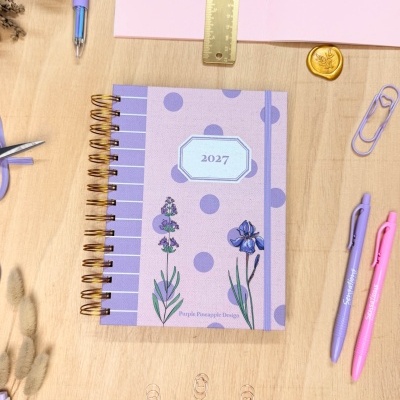 Caderno lilás com flores e bolinhas, texto 2027 e Purple Pineapple Design sobre mesa de madeira com material de escritório