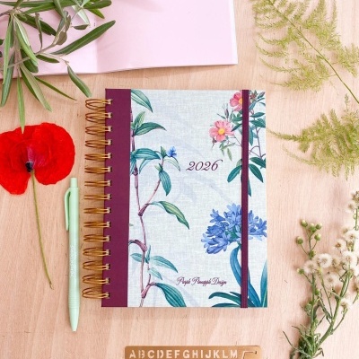 Agenda 2026 com capa floral, caneta e estêncil sobre mesa de madeira com flores