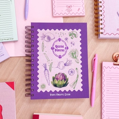 Caderno roxo e lilás com desenhos de legumes e texto RECETAS PERFEITAS rodeado por outros cadernos e canetas coloridas