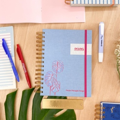 Cadernos de argolas douradas com capas azul, azul escuro e listrada, e inscrições '2026' e 'Purple Pineapple Designs'.