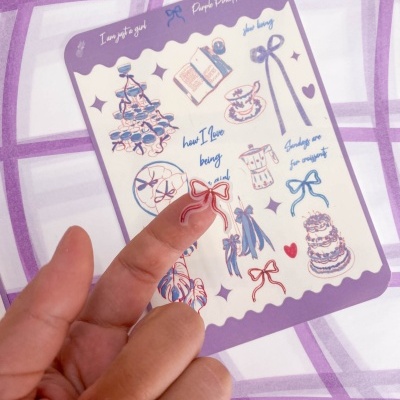 Folha de autocolantes com desenhos de lacinhos, vestidos, bolos e chávenas em cores azul, rosa e roxo.