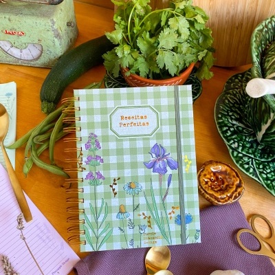 Caderno de receitas com capa verde xadrez e flores sobre mesa com legumes e utensílios