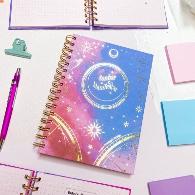 Caderno com capa azul e rosa com texto Sonhar é Manifestar e padrões cósmicos dourados