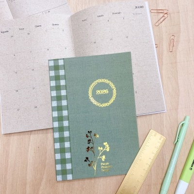 Agenda verde 2026 com padrão xadrez e flores, aberta com calendário mensal, sobre mesa de madeira com canetas e régua