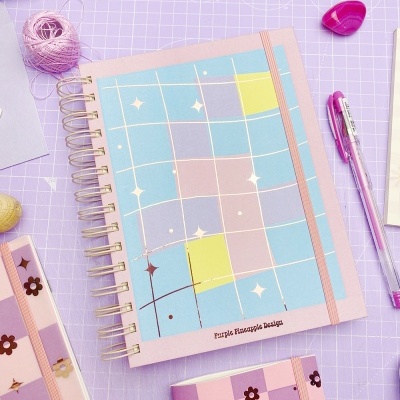 Caderno quadriculado colorido com texto Purple Pineapple Design sobre mesa lilás com acessórios de escritório